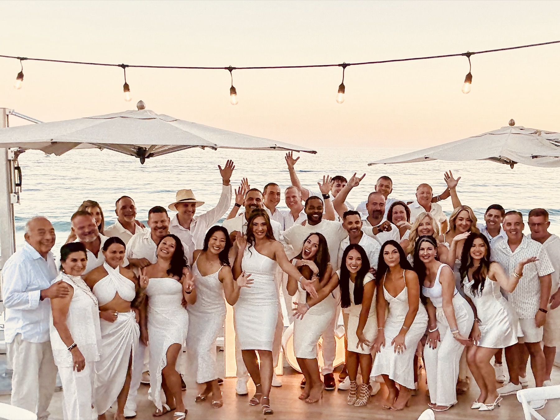 The whole crew in white, arms up at sunset on the beach deck